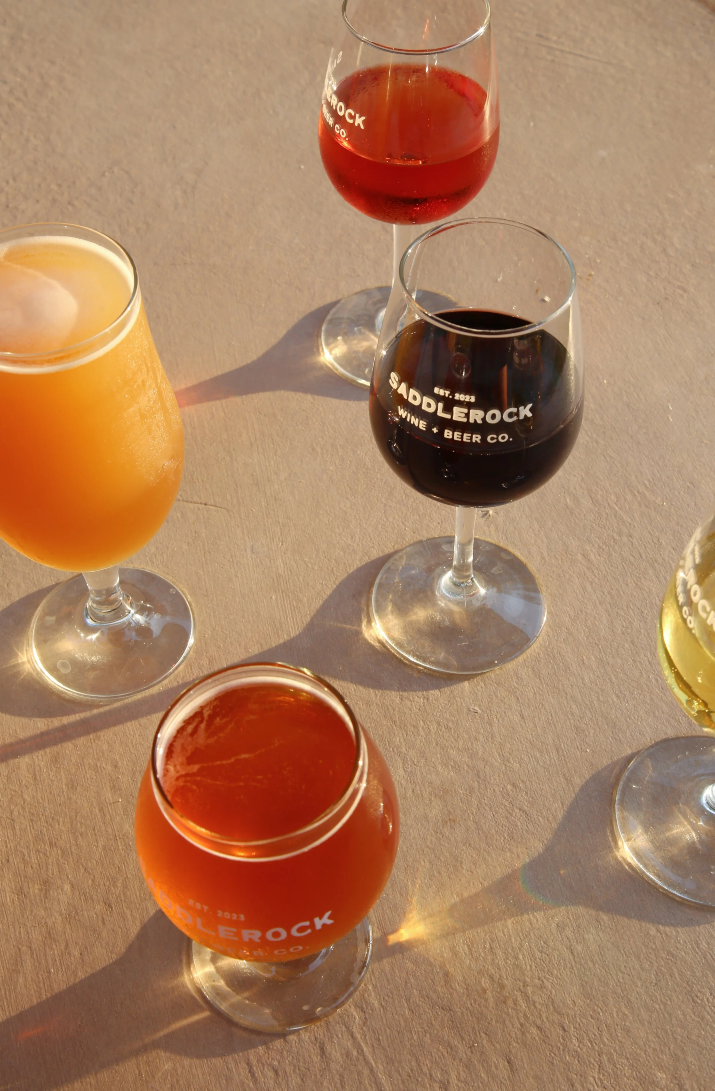A variety of colorful cocktails and wine glasses on a wooden surface, with some glasses labeled 'SADDLEROCK WINE + BEER CO.'