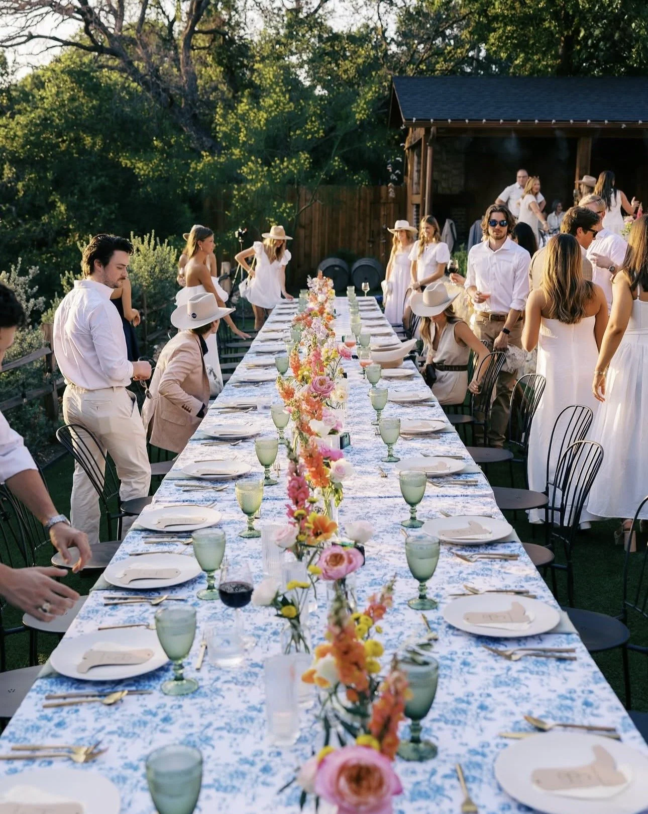 Outdoor celebration with a long dining table decorated with colorful flower arrangements, set with plates, glasses, and utensils. People dressed in white and light-colored clothing are mingling and preparing for the event, with a wooden structure and lush trees in the background.