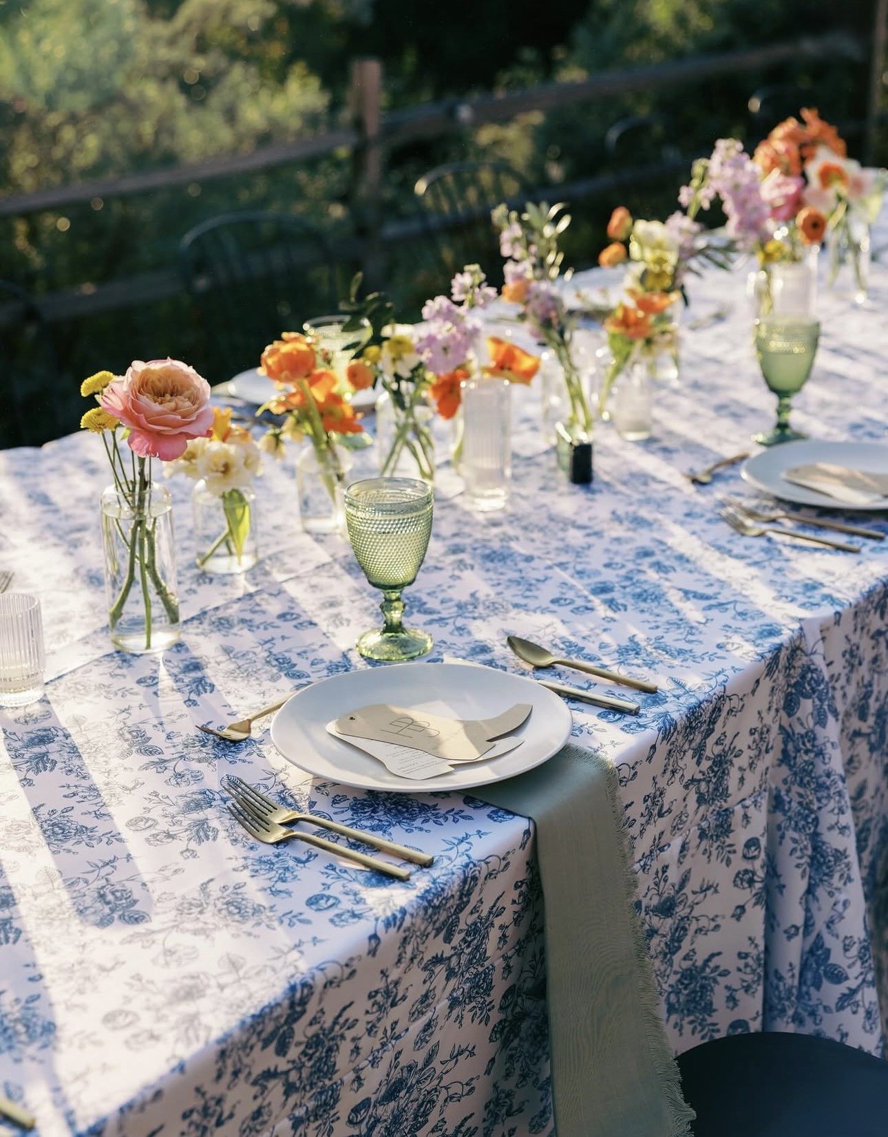 A close-up view of a beautifully set outdoor dining table covered with a white tablecloth that has blue floral patterns. The table is decorated with small glass vases holding colorful flowers, including roses and other blooms. Elegant glassware, plates, and gold-colored cutlery are arranged neatly for a meal, with one of the plates holding a small envelope or card.