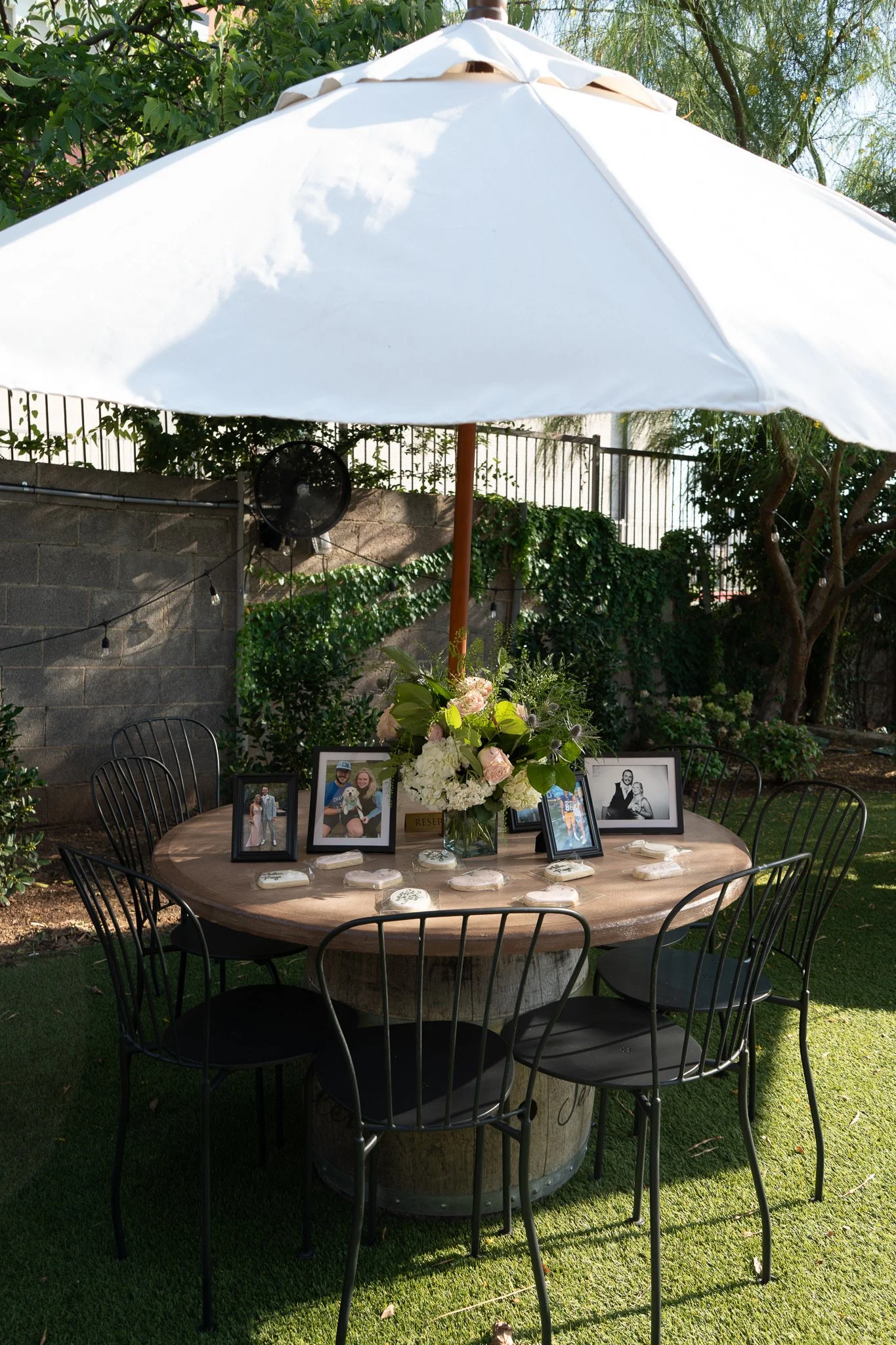 Outdoor gathering table with framed photographs and a floral centerpiece under a large white umbrella, yard with trees and a brick wall in the background.