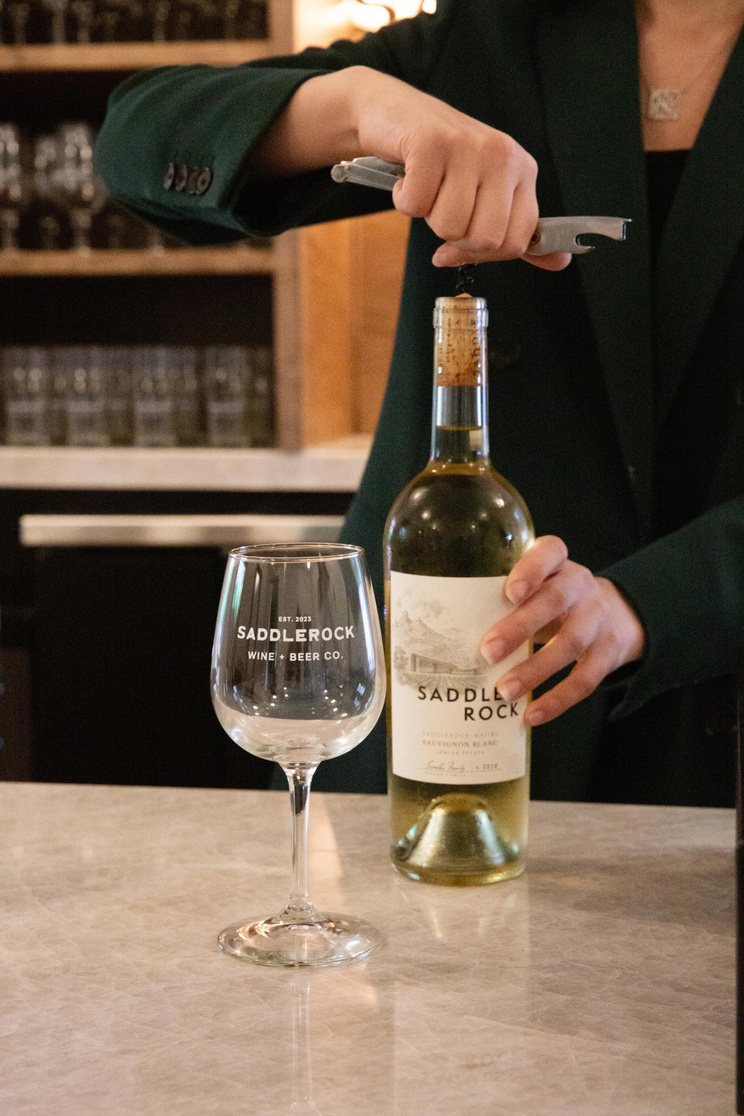 A person is opening a bottle of Saddle Rock Sauvignon Blanc wine at a bar or restaurant. In the foreground, there is an empty wine glass with the words "SADDLE ROCK WINE + BEER CO." printed on it. The background features a wooden shelf with bottles and glasses.
