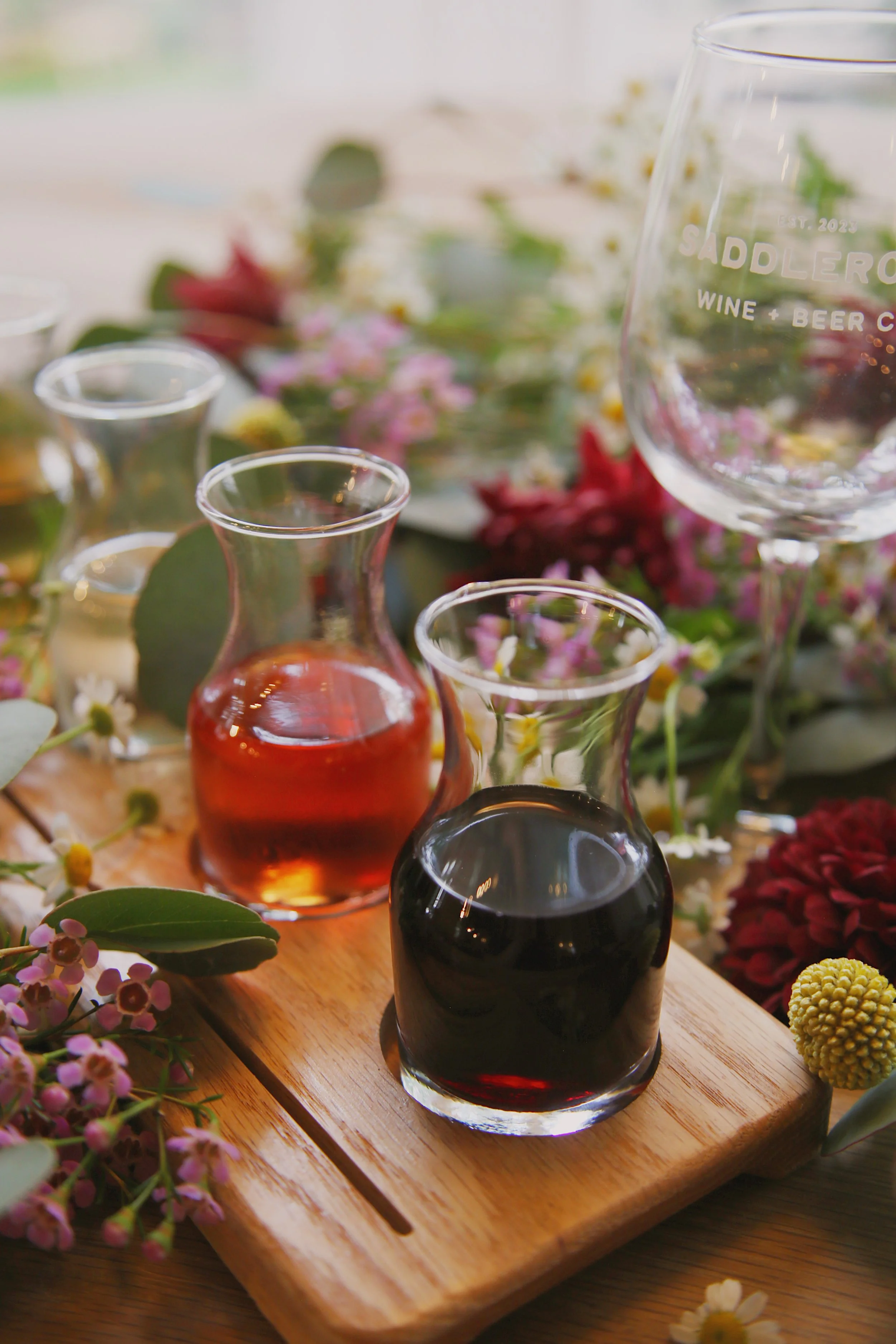 Variety of glass bottles with colorful liquids on wooden tray surrounded by flowers.