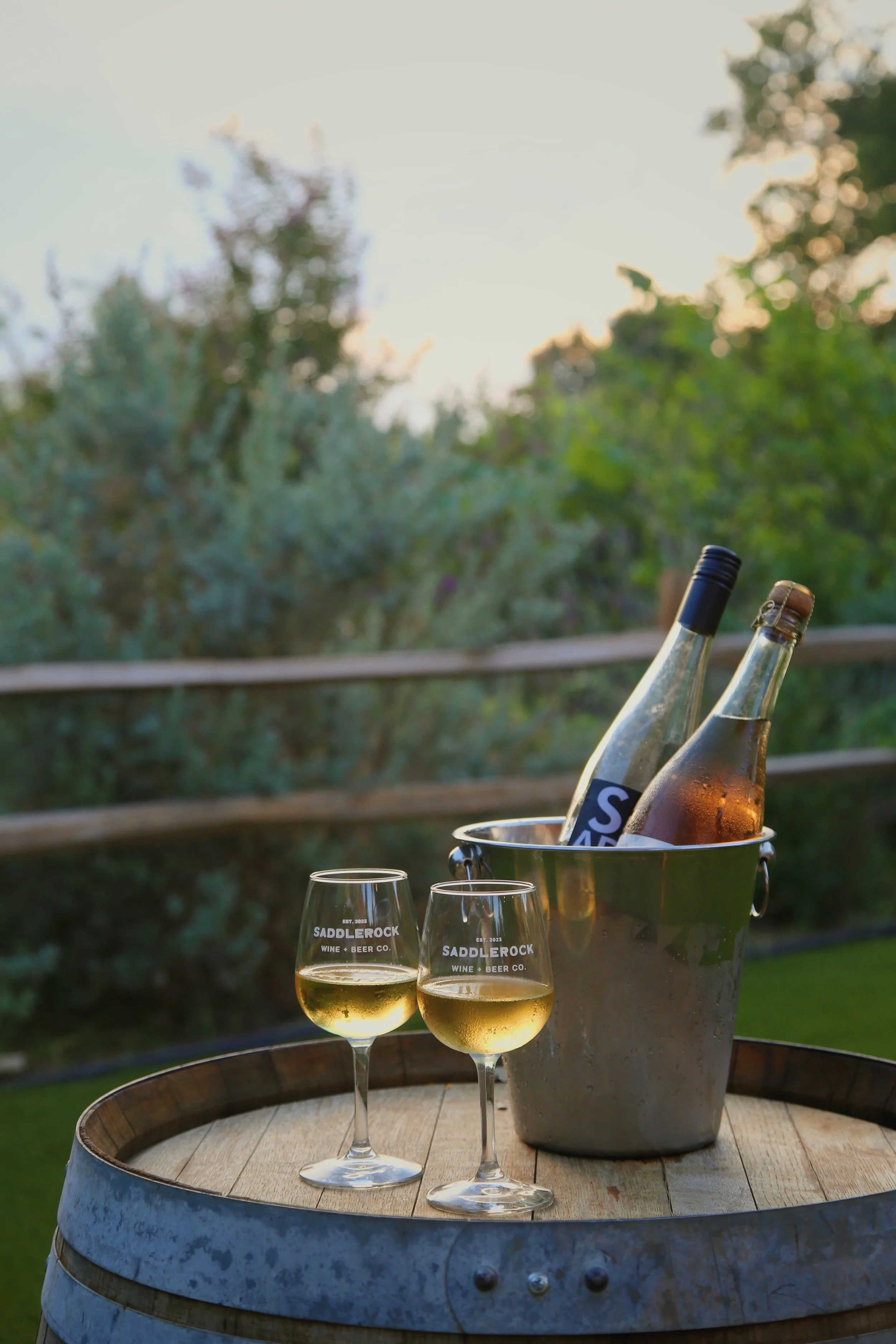 Two glasses of white wine on a wooden barrel table, with a wine cooler, wine bottles, and glasses in the background, outdoors during sunset.