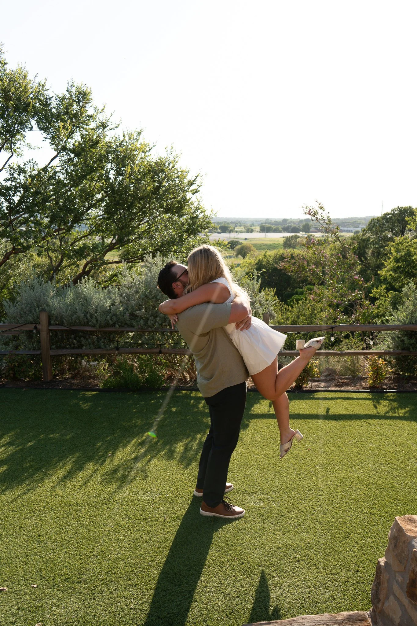 A man is holding a woman in his arms while dancing on a grassy lawn with trees and open fields in the background. The woman is wearing a white dress and heels, and the man is wearing dark pants, a light shirt, and brown shoes. The sun is shining, casting shadows on the ground.