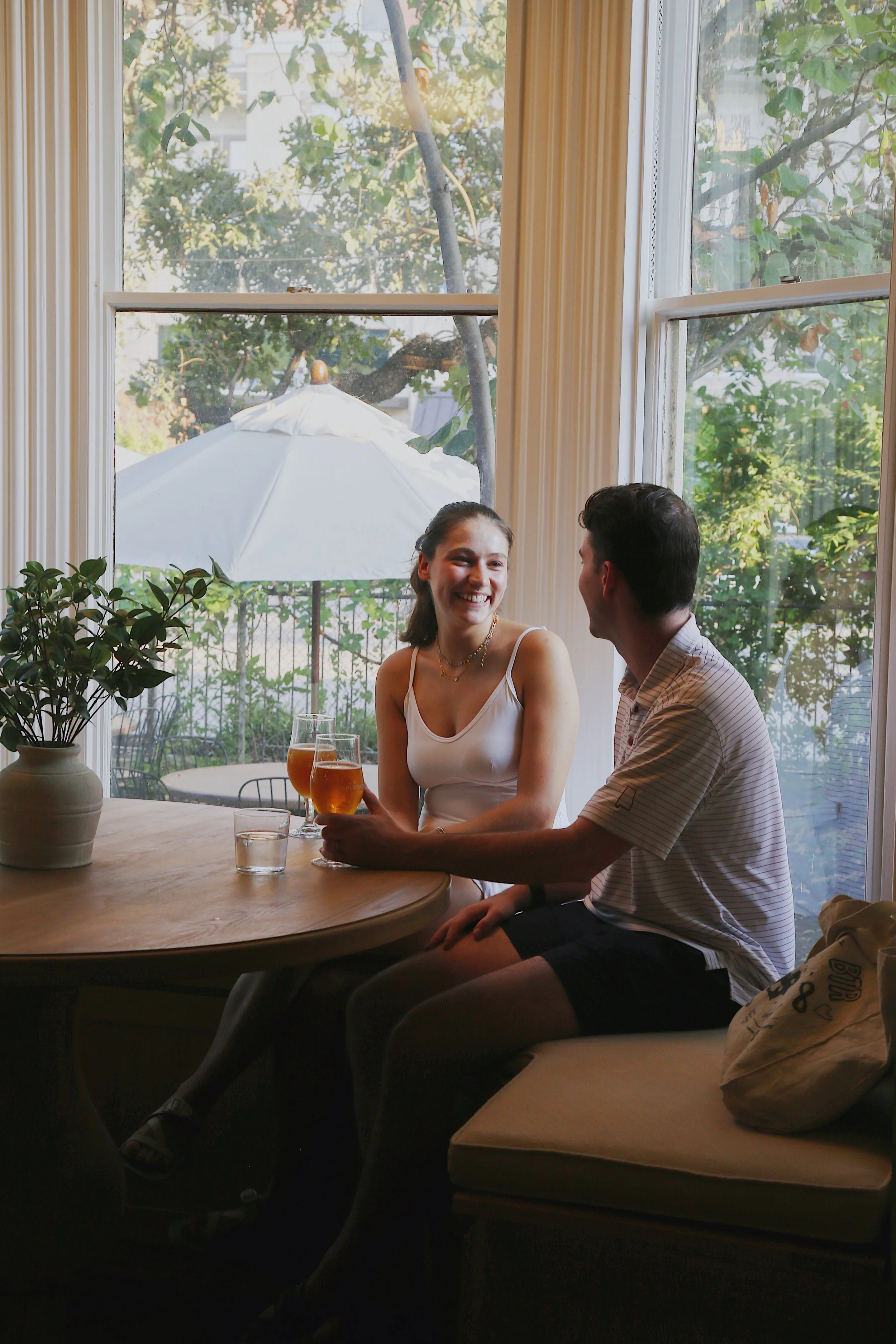 A young woman and man sitting at a round dining table inside a restaurant, smiling and talking with each other, with three glasses of beer in front of them, large windows with view of a patio with a white umbrella and green trees outside.