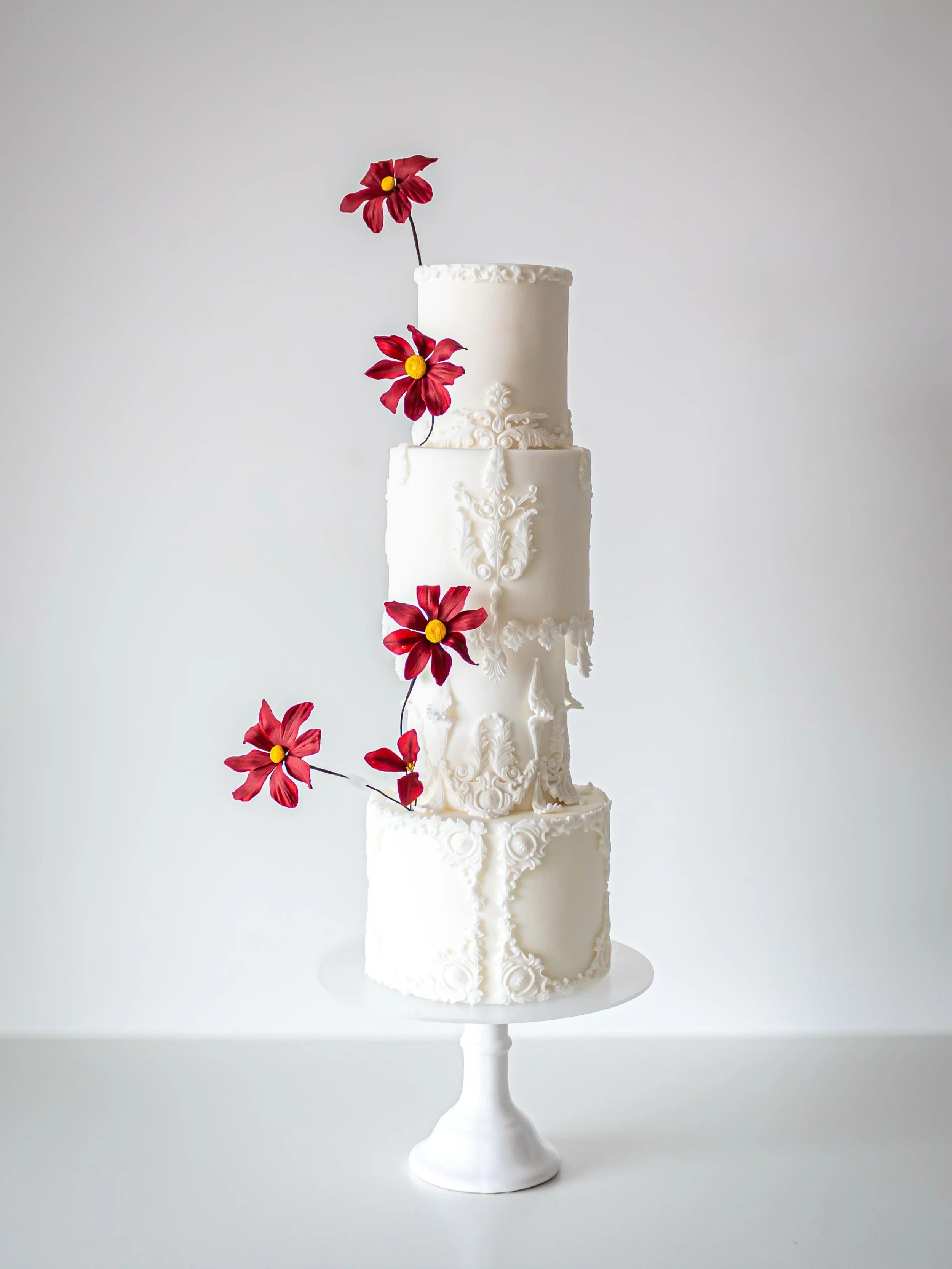 A tall, elegant white wedding cake with intricate decorative piping, topped with red flowers, displayed on a white cake stand against a plain light background.