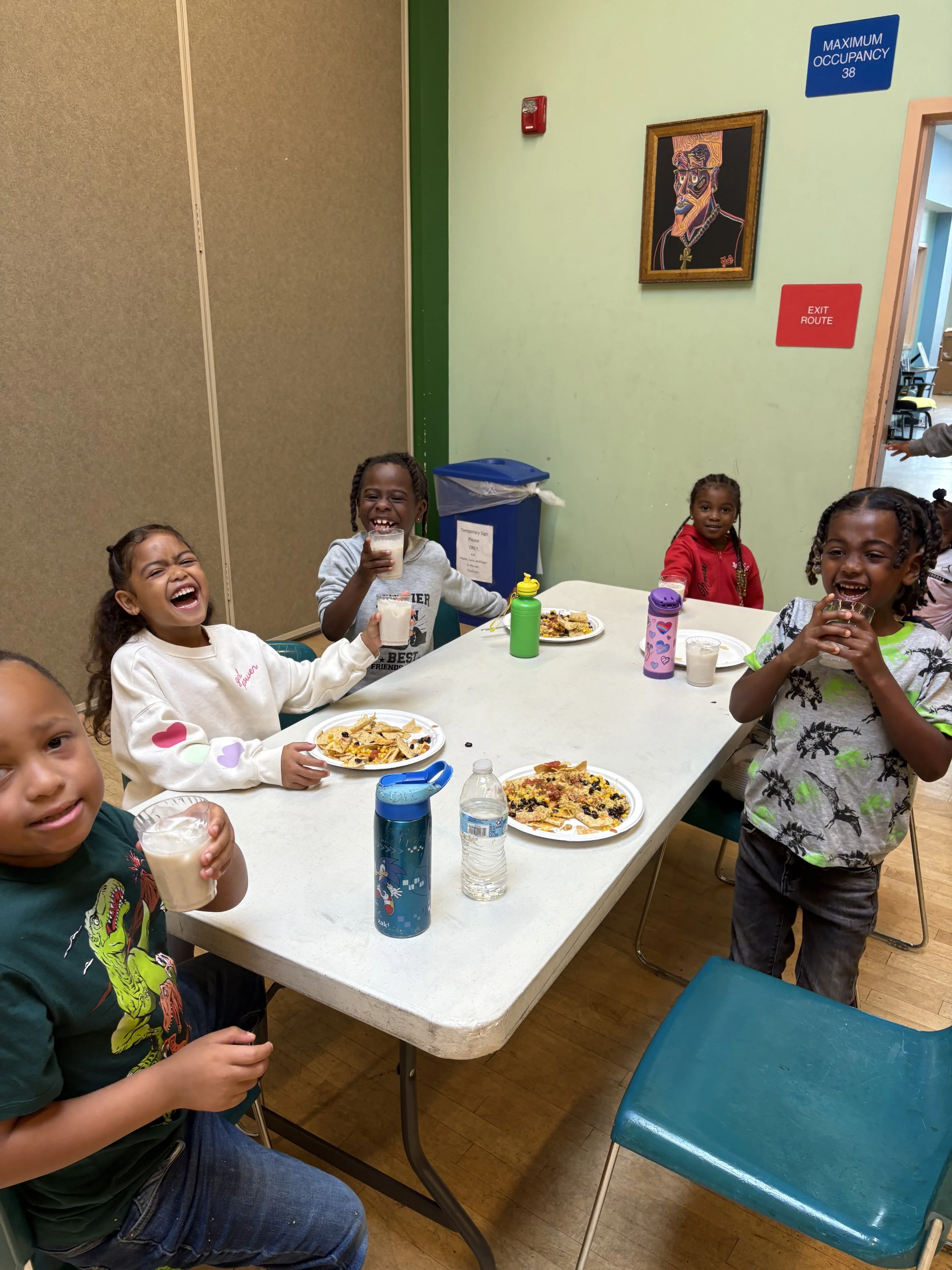 Group of five children sitting around a table with pizza slices and drinks, smiling and laughing in a colorful indoor setting.