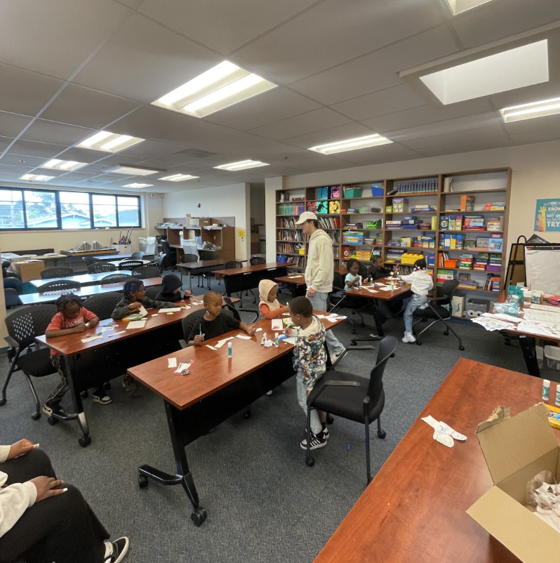 Children and adults gathered around a table engaged in arts and crafts activities in an indoor community center. The table is covered with art supplies, including glue, paint, and decorative items.