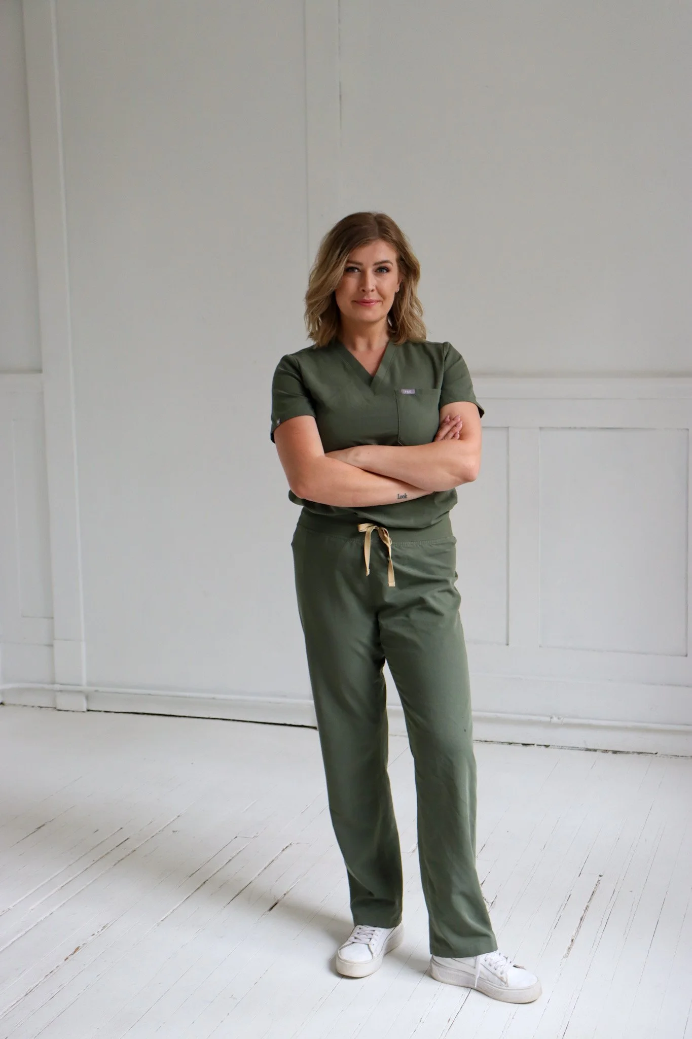 A woman with shoulder-length blonde hair dressed in green medical scrubs and white sneakers, standing with her arms crossed in a minimal white room.