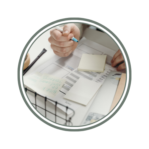 Close-up of a person's hand writing on a piece of paper with sticky notes and a pen on a desk covered with financial documents.