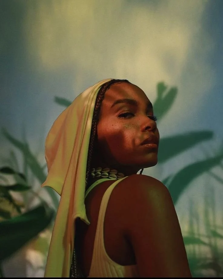 A woman with braids and a headscarf looking over her shoulder in front of a blurred background of green leafy plants.