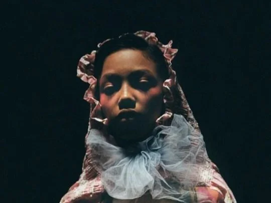 A woman with a serious expression, wearing a lace headscarf with ruffles and a large, sheer bow around her neck, against a dark background.