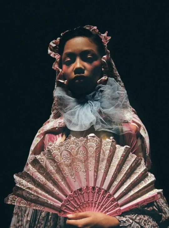 A portrait of a young girl with dark skin wearing historical clothing, holding a pink and gold fan, with a dark background.