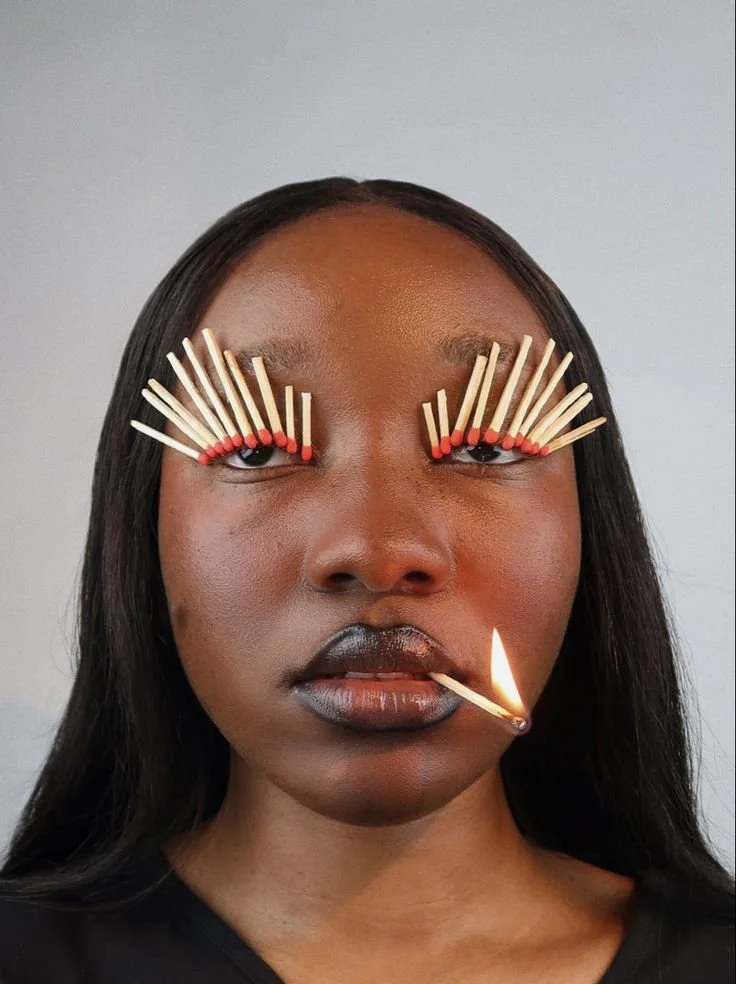 A woman with eyelash extensions made of matches, with some lit matches in her mouth.