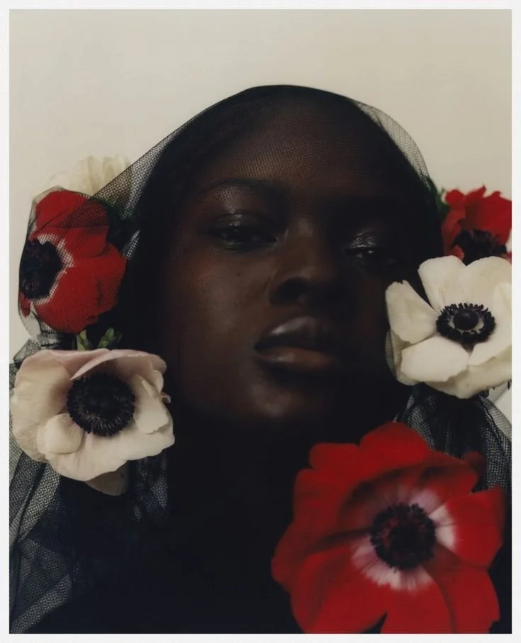 A close-up portrait of a woman with dark skin, surrounded by white, red, and black flowers and a black mesh veil.