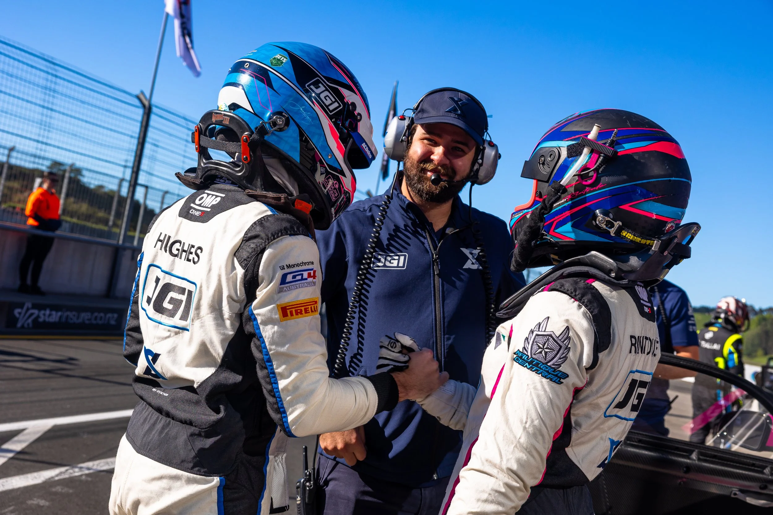 Two race car drivers in racing suits and helmets shaking hands and talking with a crew member in a blue jacket and headset at the racing track during daytime.