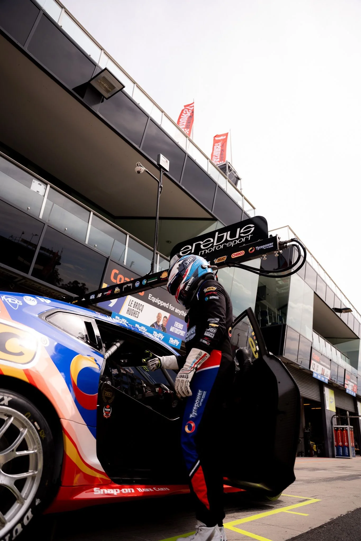 A race car driver in racing gear and helmet standing next to a racing car in the pit lane of a motorsport circuit.