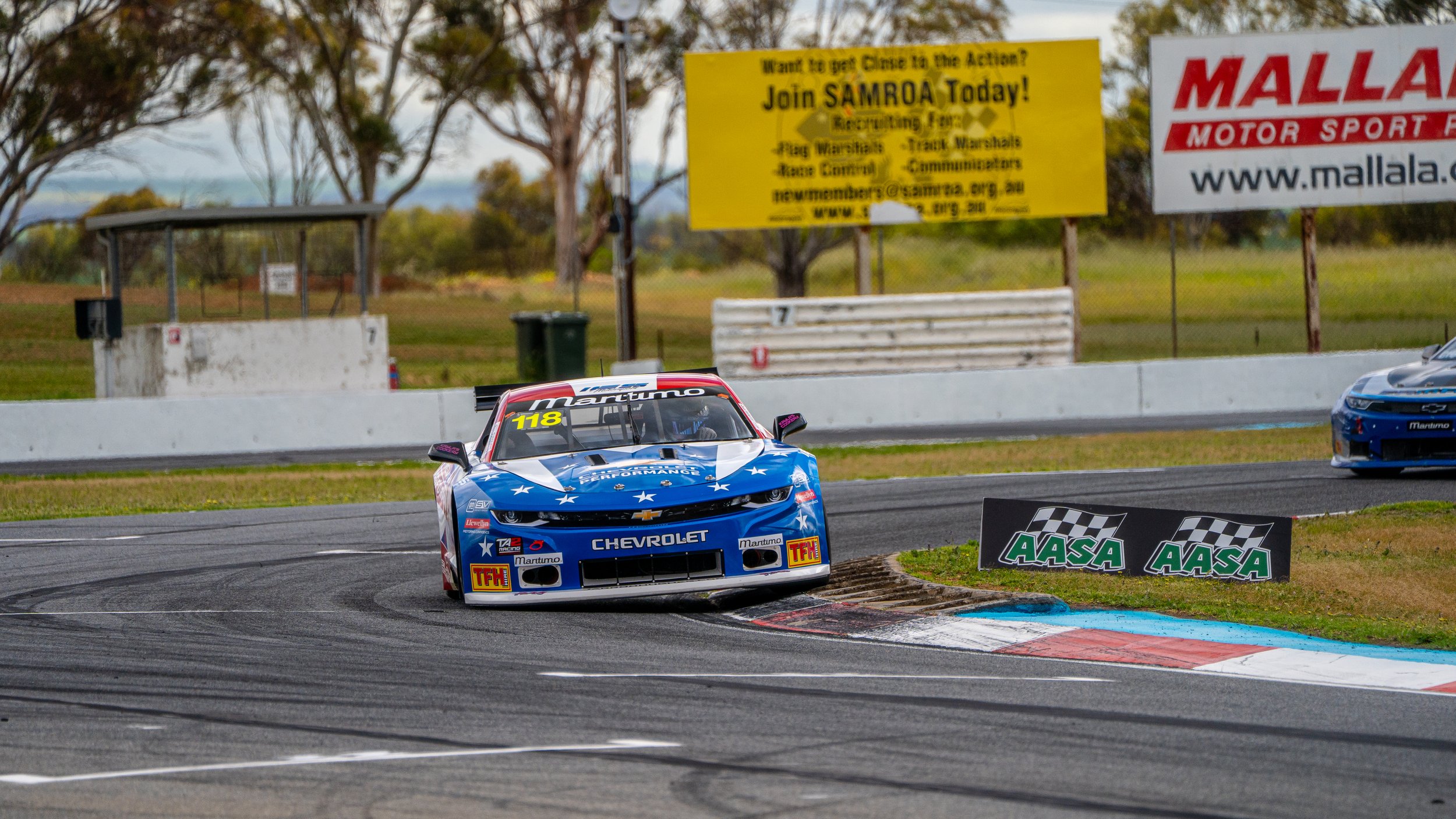 A blue Chevrolet race car with the number 118 on the windshield speeds around a race track, with a black and white checkered flag sign nearby. The background shows trees and advertising banners, including one for MALLALA MOTOR SPORT.