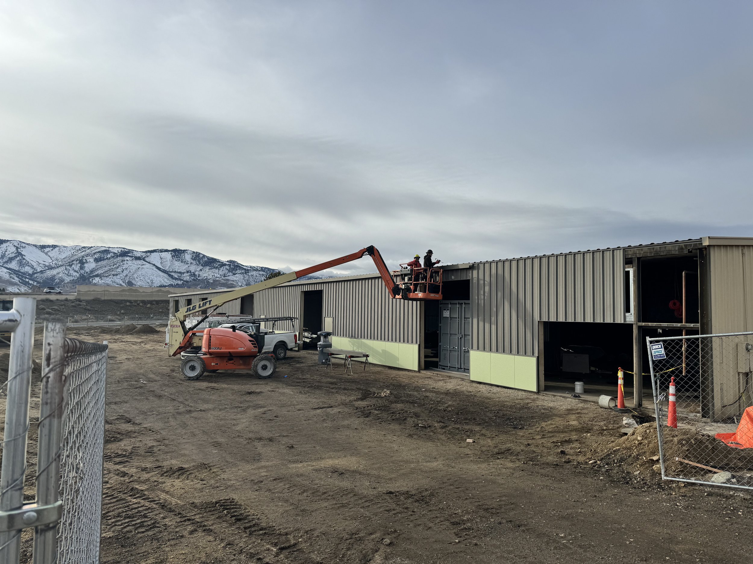 Construction site with partially built metal structure, JLG lift, and workers on the roof; mountains and overcast sky in background