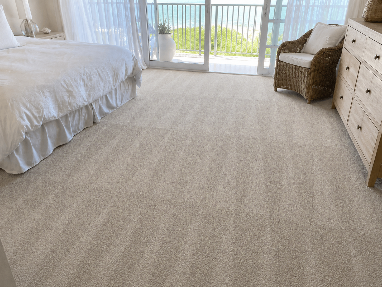 A bedroom with a large sliding glass door leading to a balcony with a view of greenery and the ocean. The room has a bed with white bedding, a wicker chair with white cushions, a white dresser, a nightstand with a lamp and decorative item, and beige carpet flooring.
