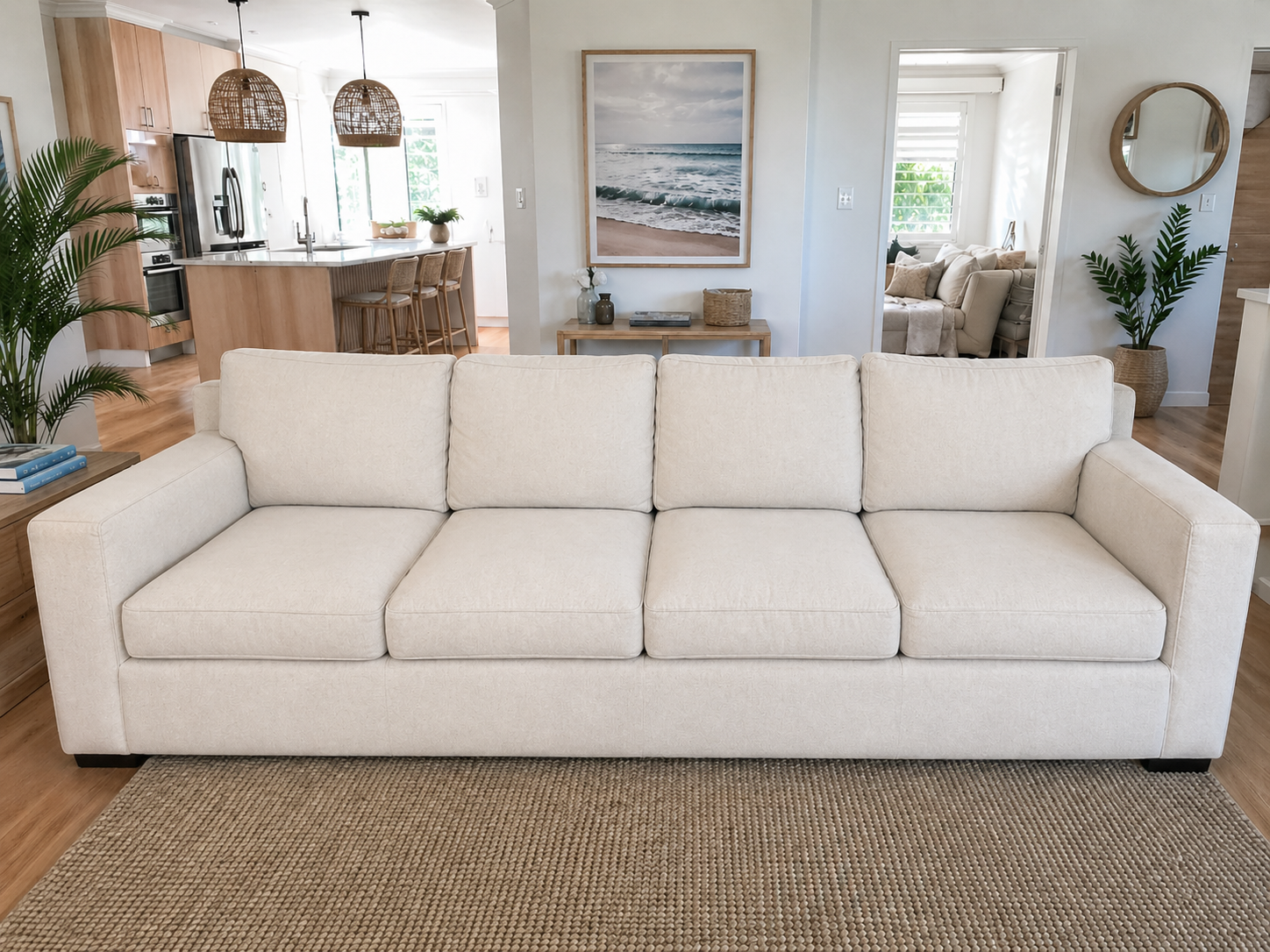 A beige sofa in a living room with a wood floor, a brown rug, and houseplants, with an open kitchen and another living area in the background.