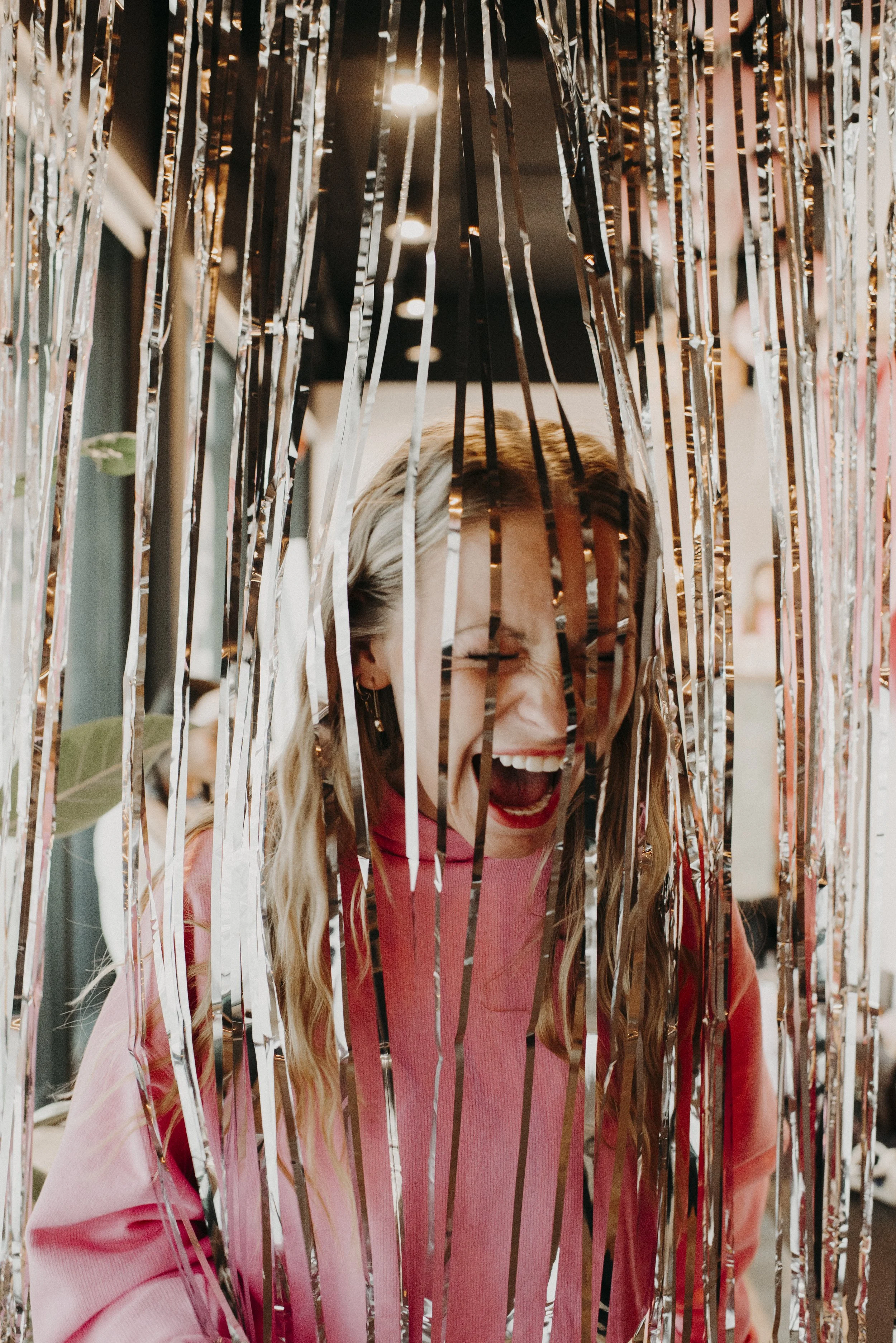 Woman with blonde hair and a pink shirt, smiling and laughing behind metallic silver and gold tinsel decorations.