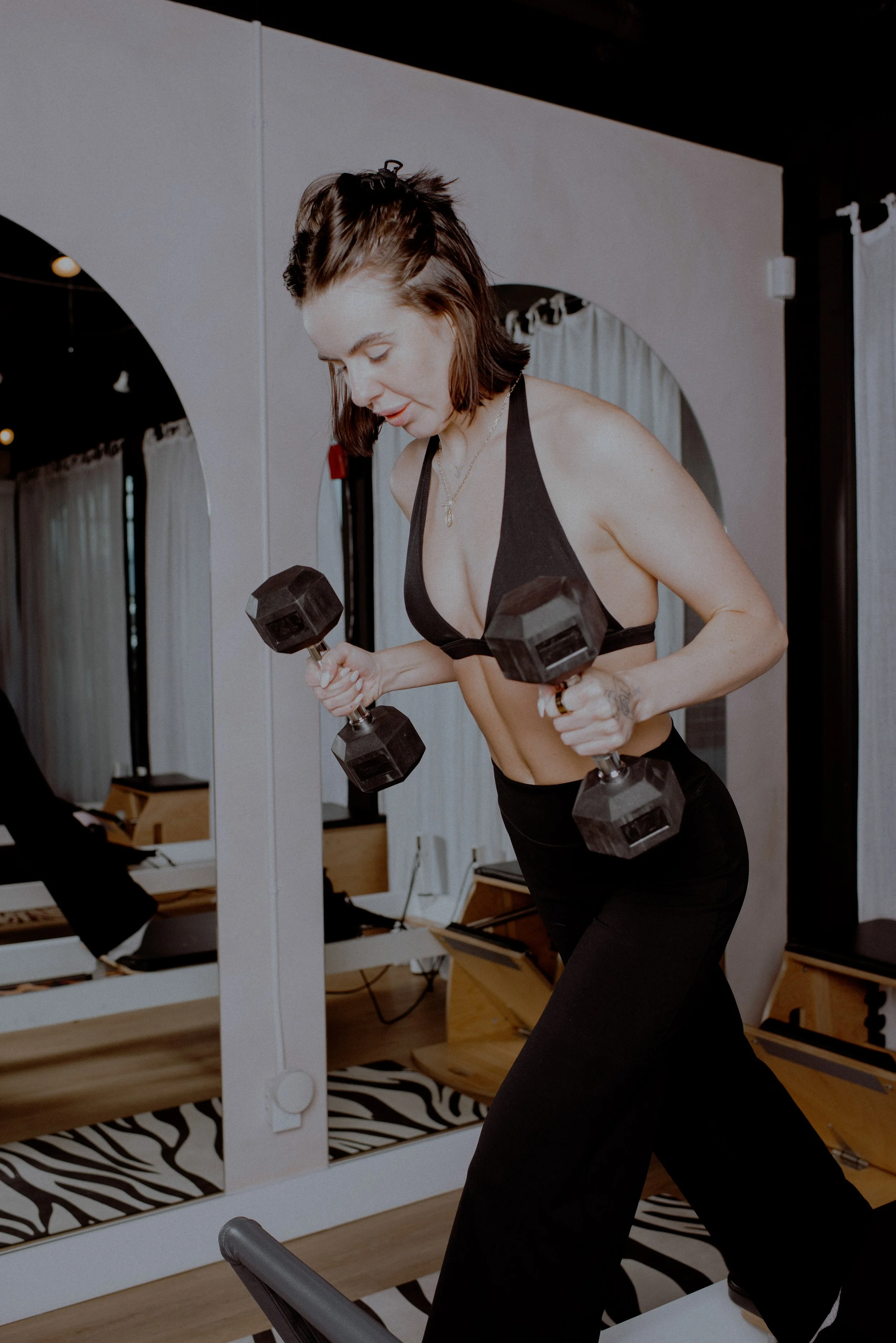 A woman in workout attire holding light green dumbbells in a fitness studio with arched mirrors and soft lighting.