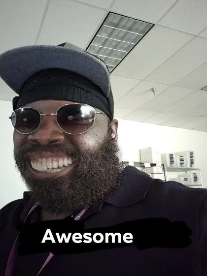 Close-up of a man with a beard wearing sunglasses, a gray cap, and a black hoodie, smiling in an office setting with fluorescent ceiling lights and filing cabinets in the background.