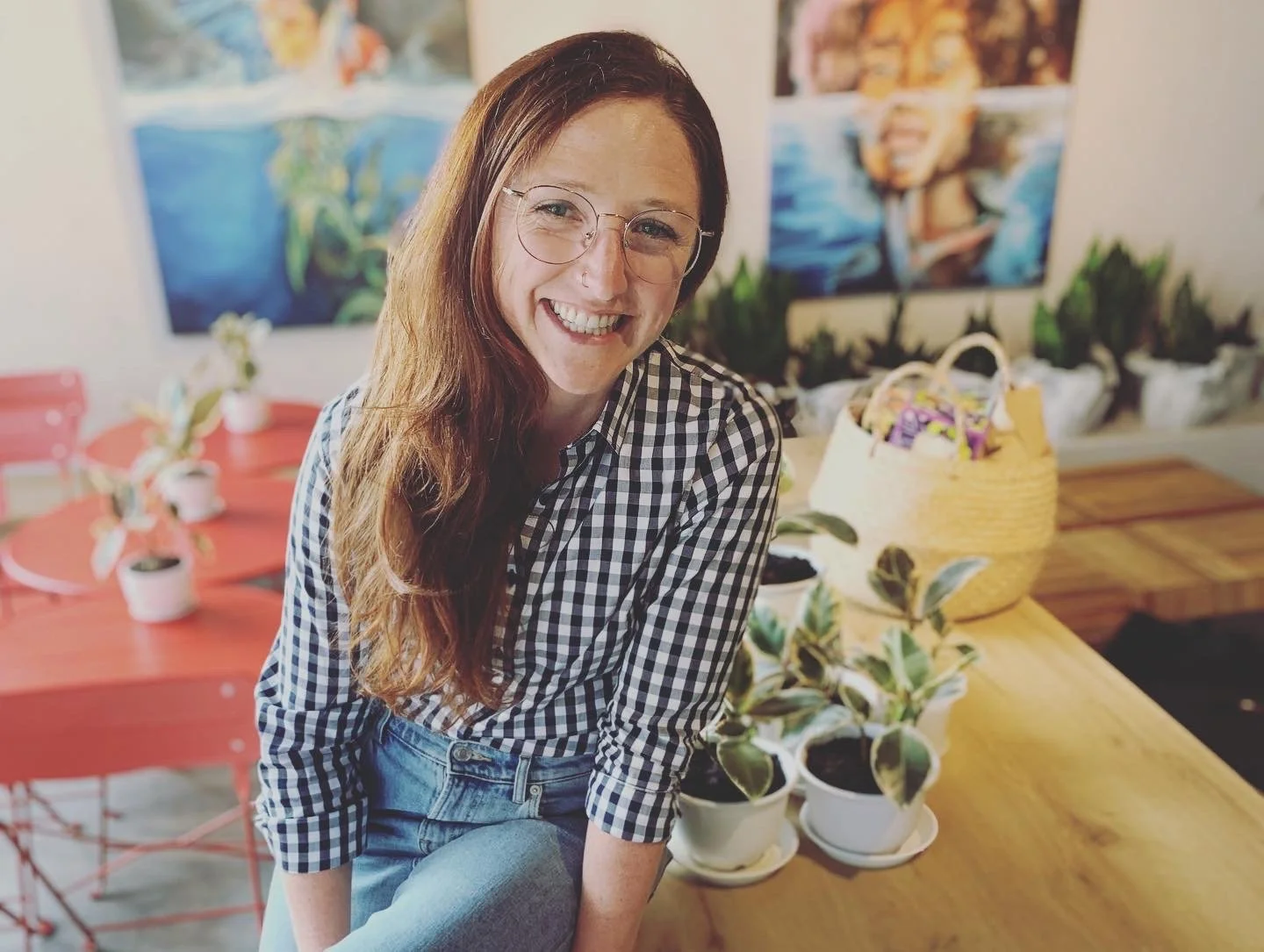 A death doula with long red hair, glasses, and a checkered shirt smiles in a cafe, surrounded by potted plants, colourful paintings on the wall, and a wooden table.