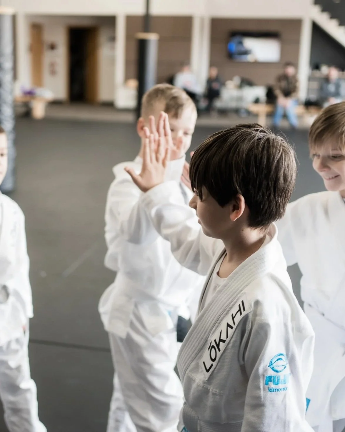 Young children practicing Brazilian Jiu-Jitsu in a martial arts gym, receiving high fives from their instructor, all wearing white gi uniforms with martial arts patches.