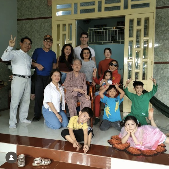 Family group photo in front of a house, with elderly woman seated in the center, children and adults around her, some making peace signs, some sitting on the floor and some standing.