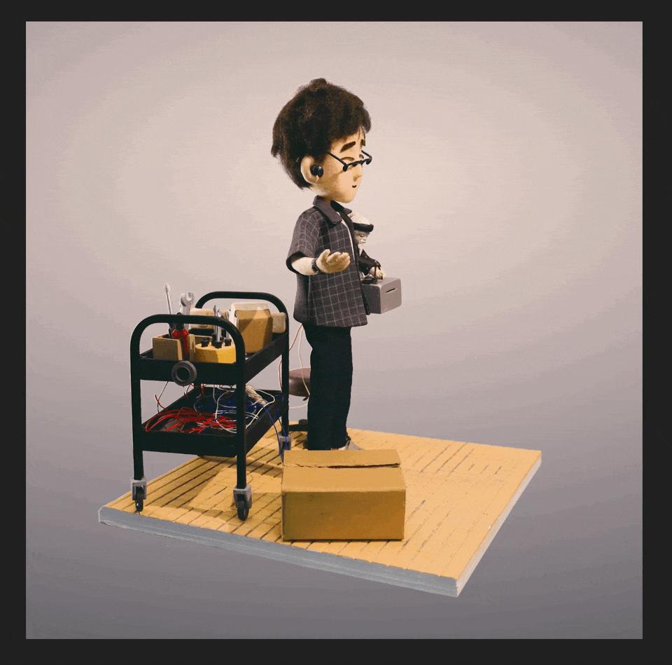 A puppet of a man with glasses and dark hair, wearing a dark shirt and pants, standing on a small wooden platform. The puppet holds a remote control and is connected to wires, with a cart full of tools and a cardboard box nearby.