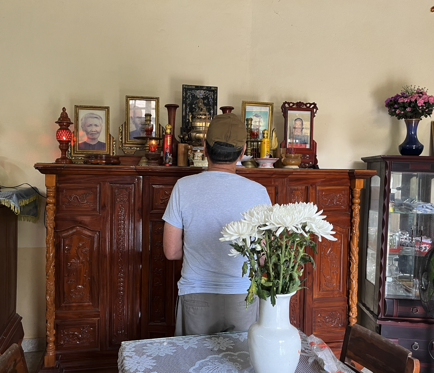 A man wearing a tan cap and a light gray T-shirt standing in front of an altar with framed photos, candles, and religious artifacts inside a home. A white vase with white flowers is on a table in the foreground.