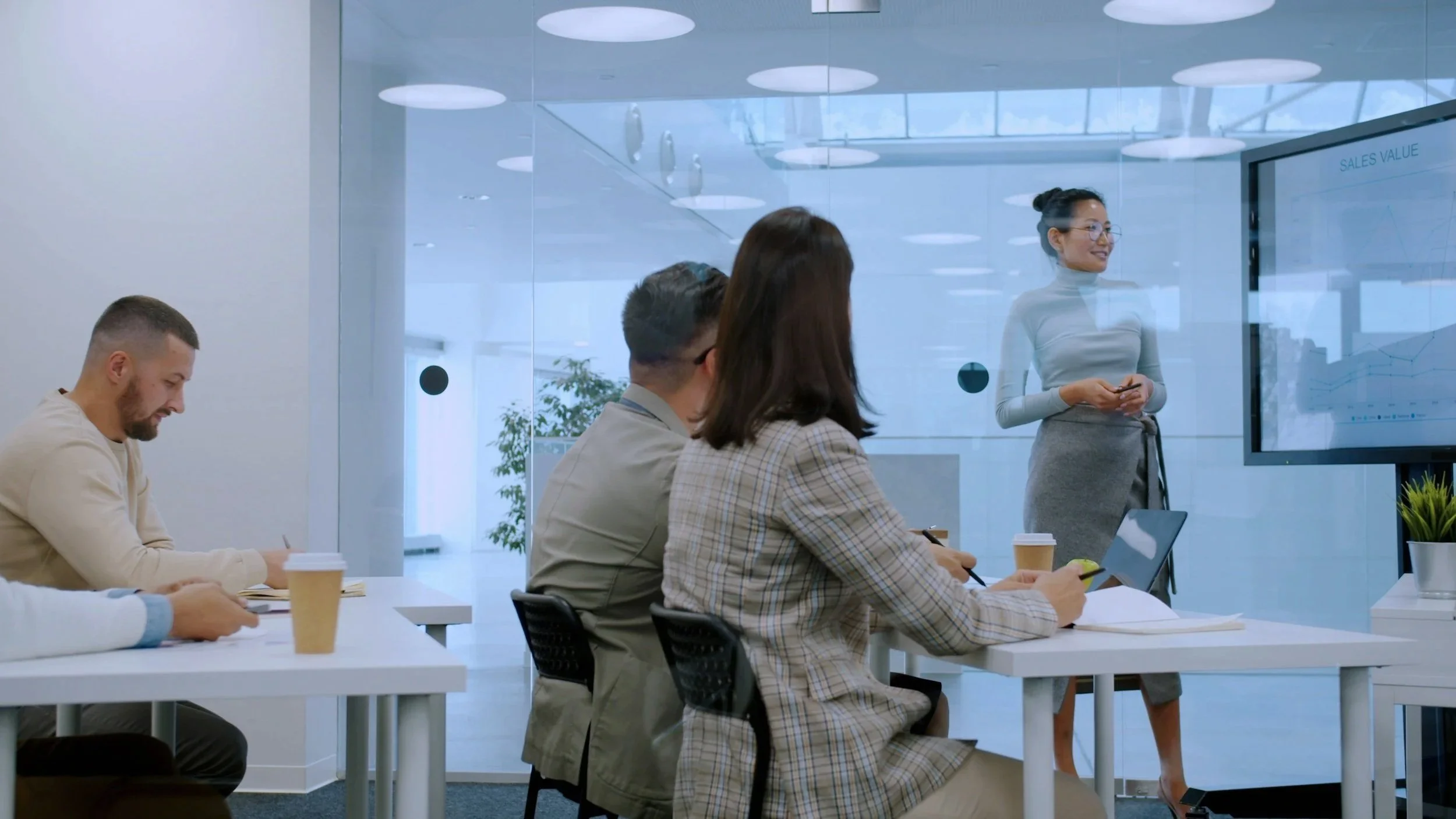 A woman standing and presenting a sales chart to a small group of people seated at tables in a modern conference room with glass walls.