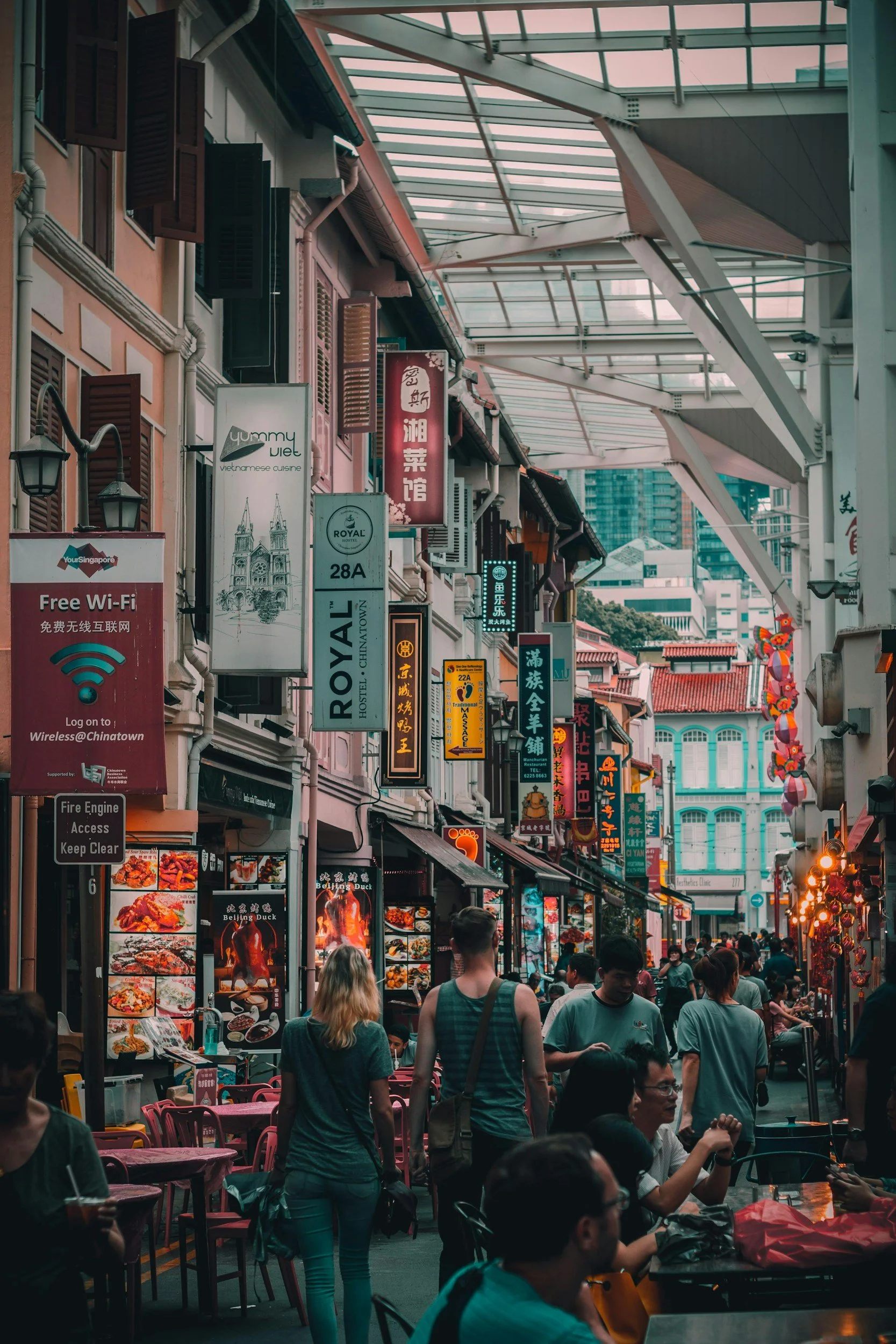 Bustling street scene in Chinatown with numerous signs in Chinese characters and English, outdoor dining with tables and chairs, people walking and sitting, restaurant menus, and a glass canopy overhead.