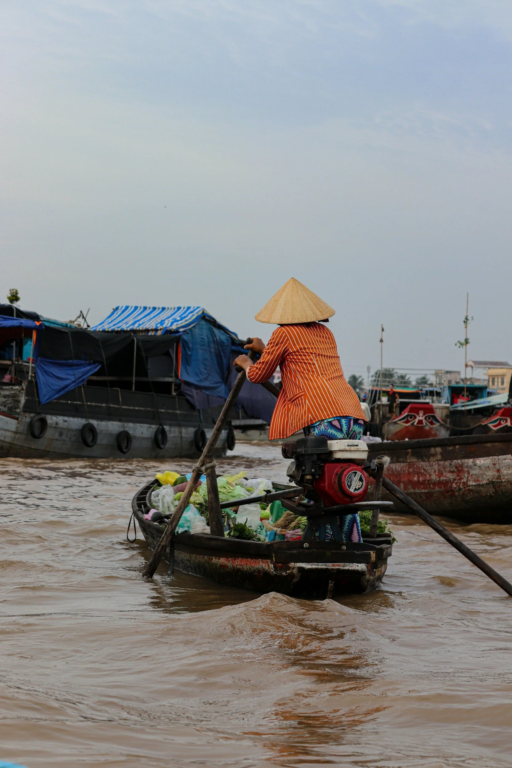A person wearing a conical Asian hat and an orange striped shirt paddles a small boat filled with vegetables on a river. Several boats are docked in the background, some with blue tarps, and the sky is overcast.