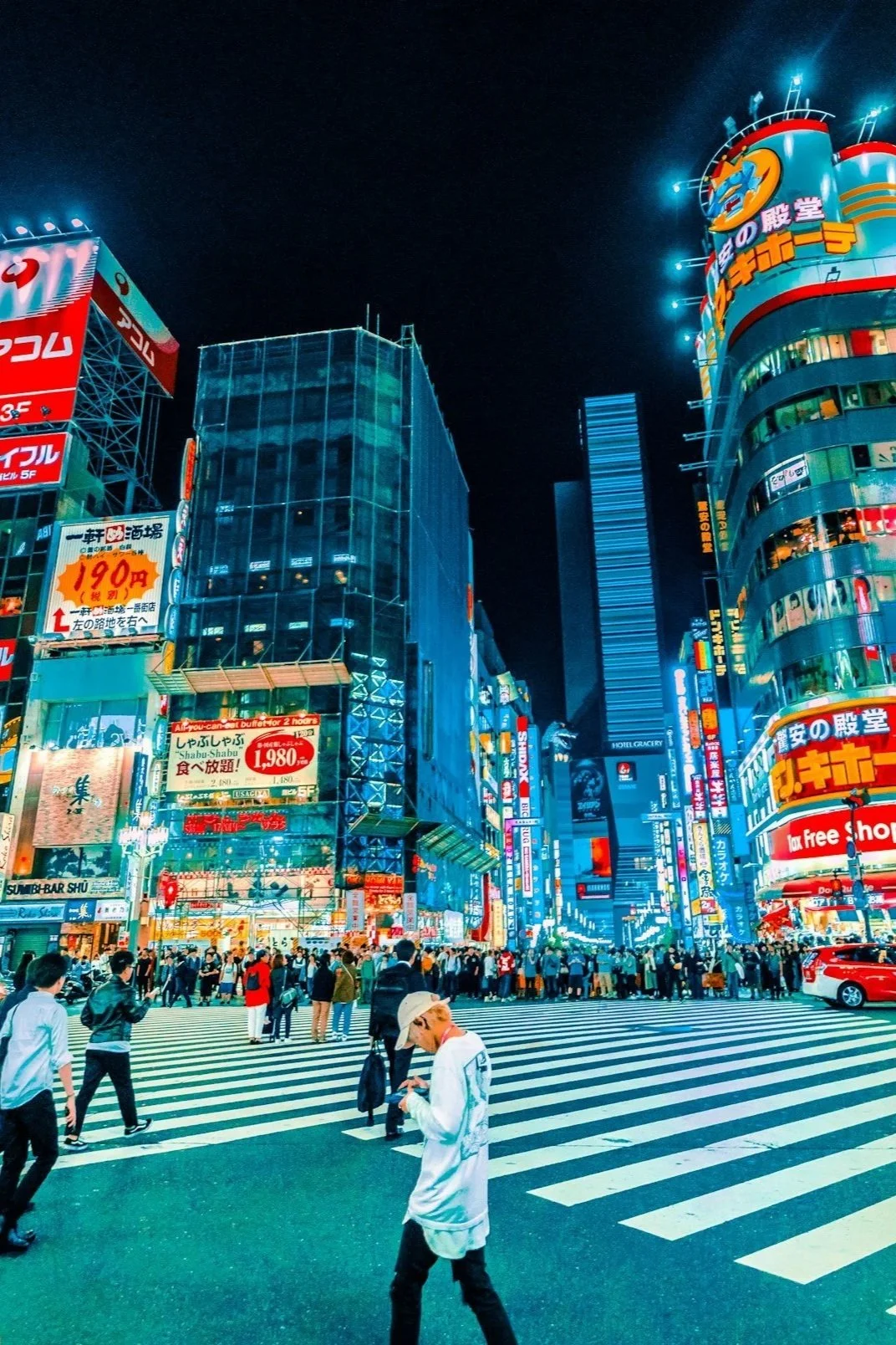 Nighttime city scene at a busy crosswalk in an urban area of Japan, with brightly lit buildings, colorful neon signs in Japanese, and pedestrians crossing the street.