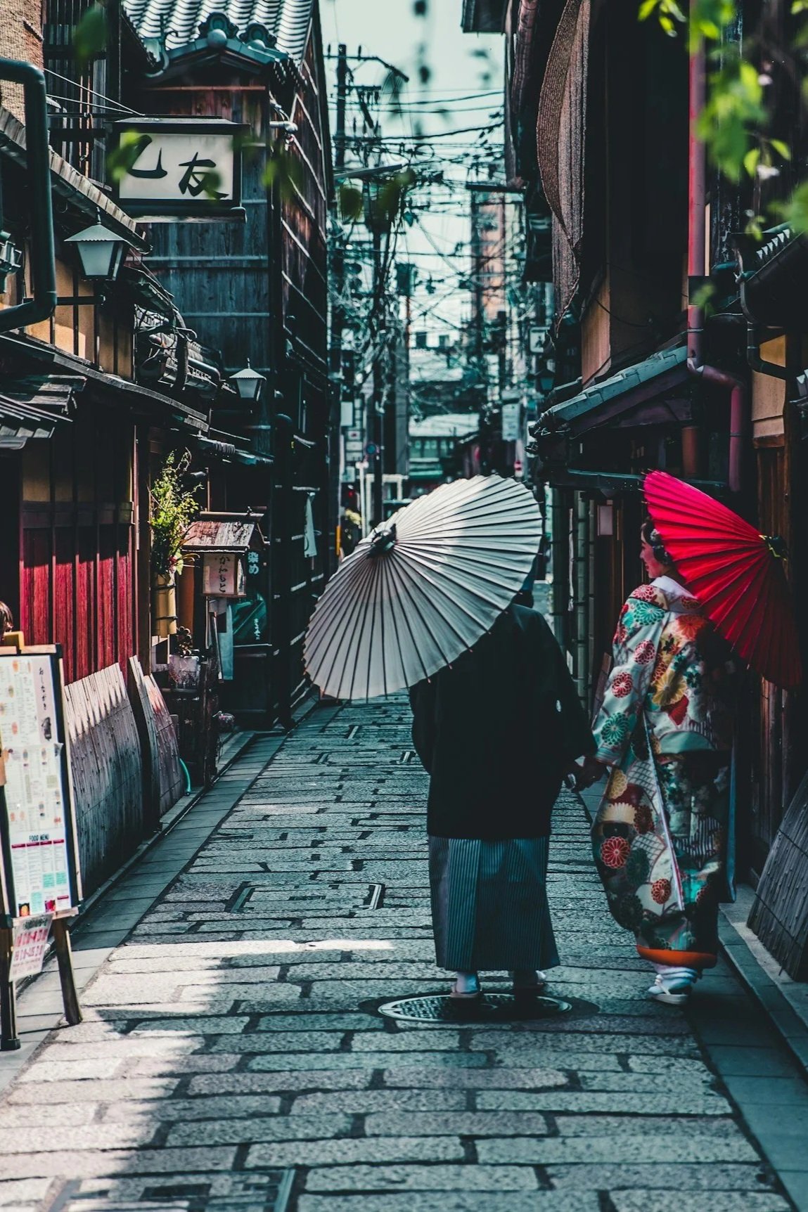 A narrow street in Japan with two women dressed in traditional kimonos, holding umbrellas, walking past shops with wooden facades.