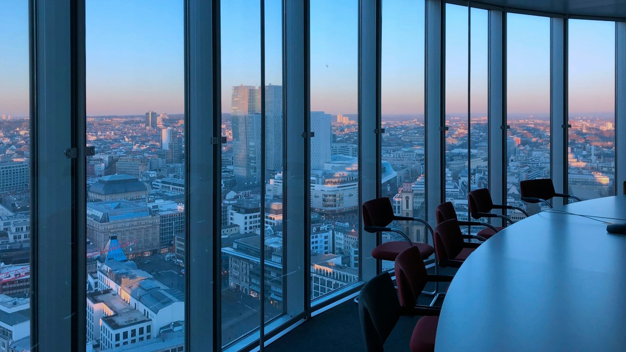 City skyline view from inside a modern conference room with floor-to-ceiling glass windows, office chairs, and a table.