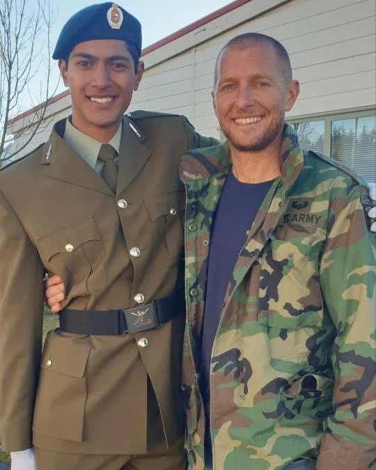 Two men smiling and standing close together outdoors, one in a military uniform with a blue beret and the other in a camouflage army jacket.