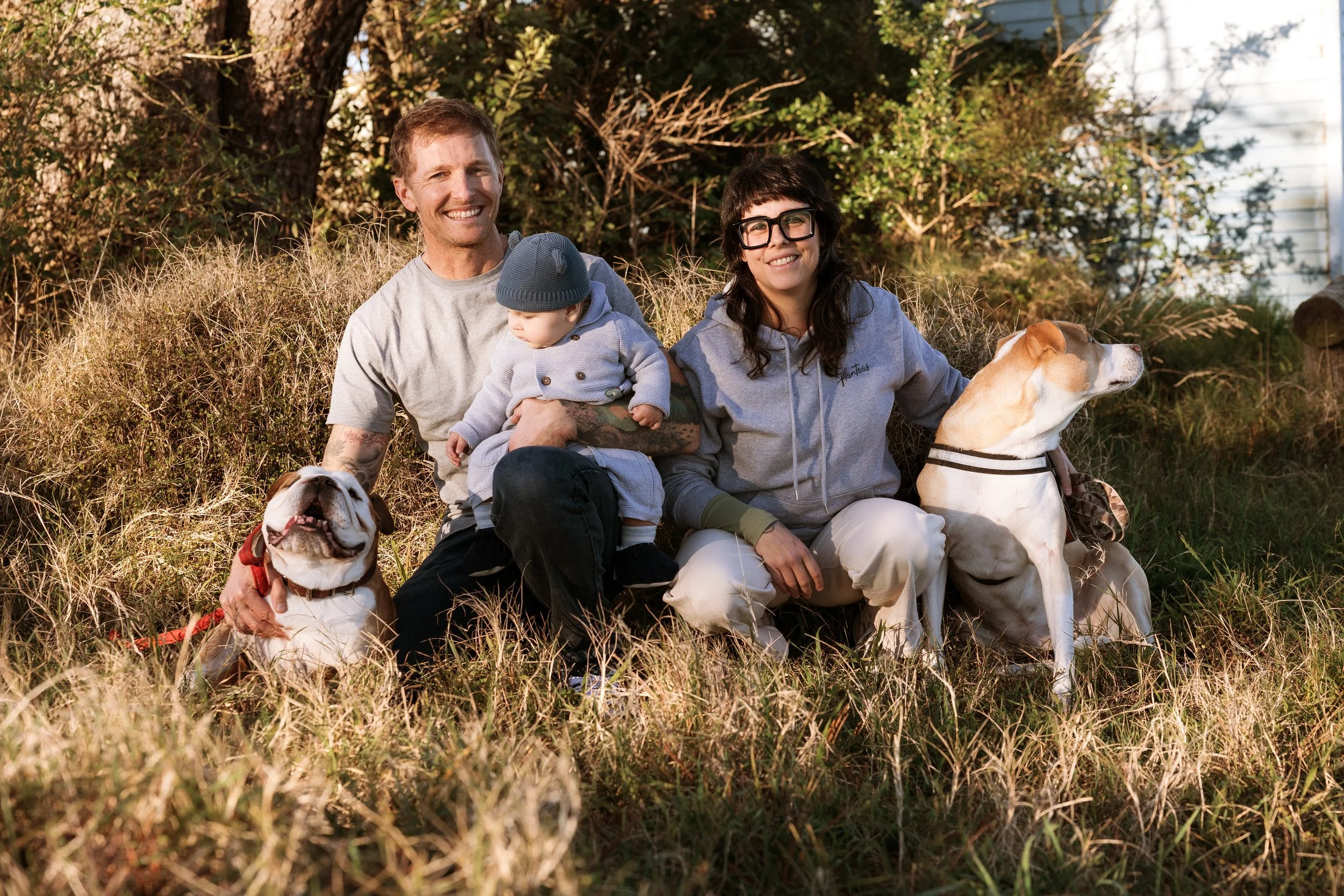A happy family with two adults, a baby, and two dogs sitting outdoors in a grassy area with trees and a house in the background.