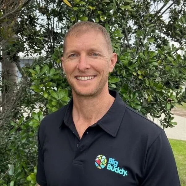 A man with short hair and a beard smiling outdoors, wearing a black polo shirt with a 'Big Buddy' logo, standing in front of green leafy bushes.