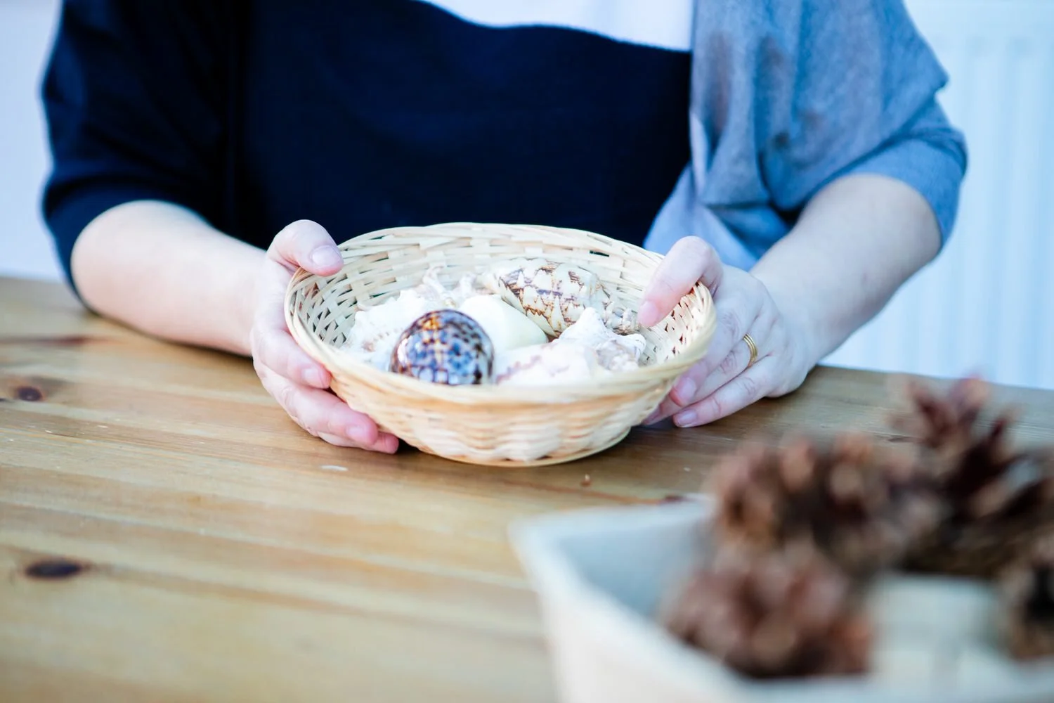 Person holding a small basket of assorted seashells on a wooden table.