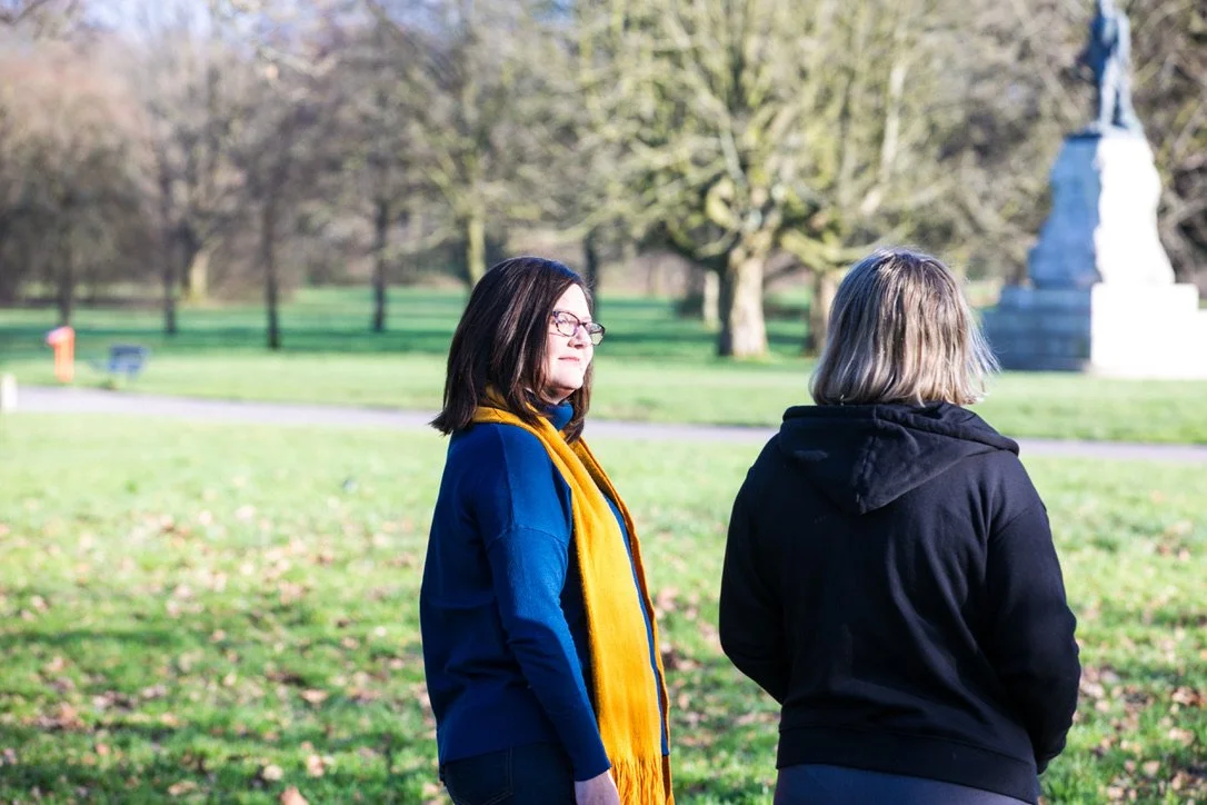 Two women standing in a park with green grass and trees, one facing left and the other facing away, with a statue visible in the background.