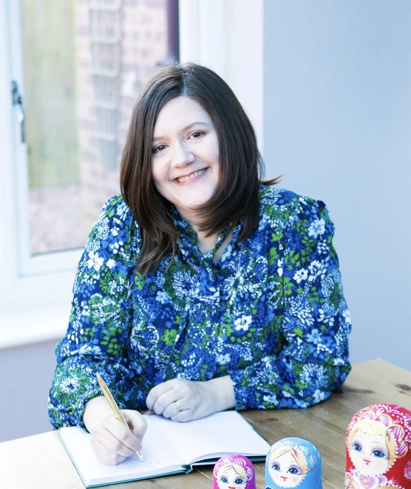 A woman with shoulder-length brown hair smiling at the camera, sitting at a table with a notebook and a gold pen, wearing a blue floral blouse, with three colorful Russian nesting dolls on the table.