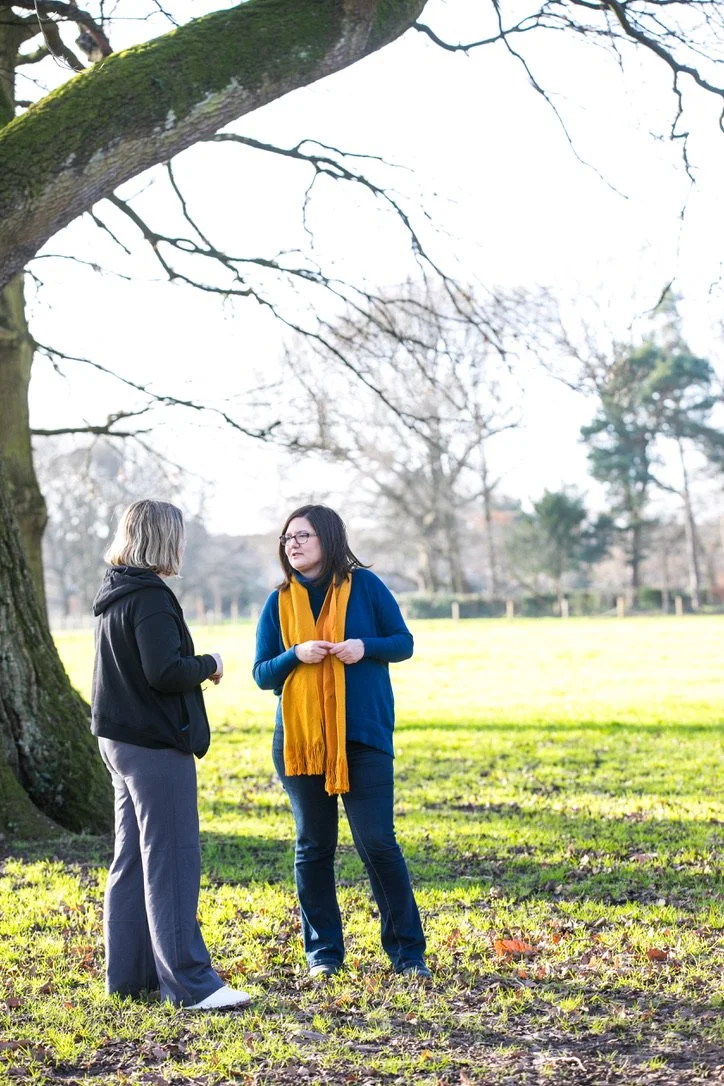 Two women standing and talking outdoors under a tree in a park with grass and leafless trees in the background.