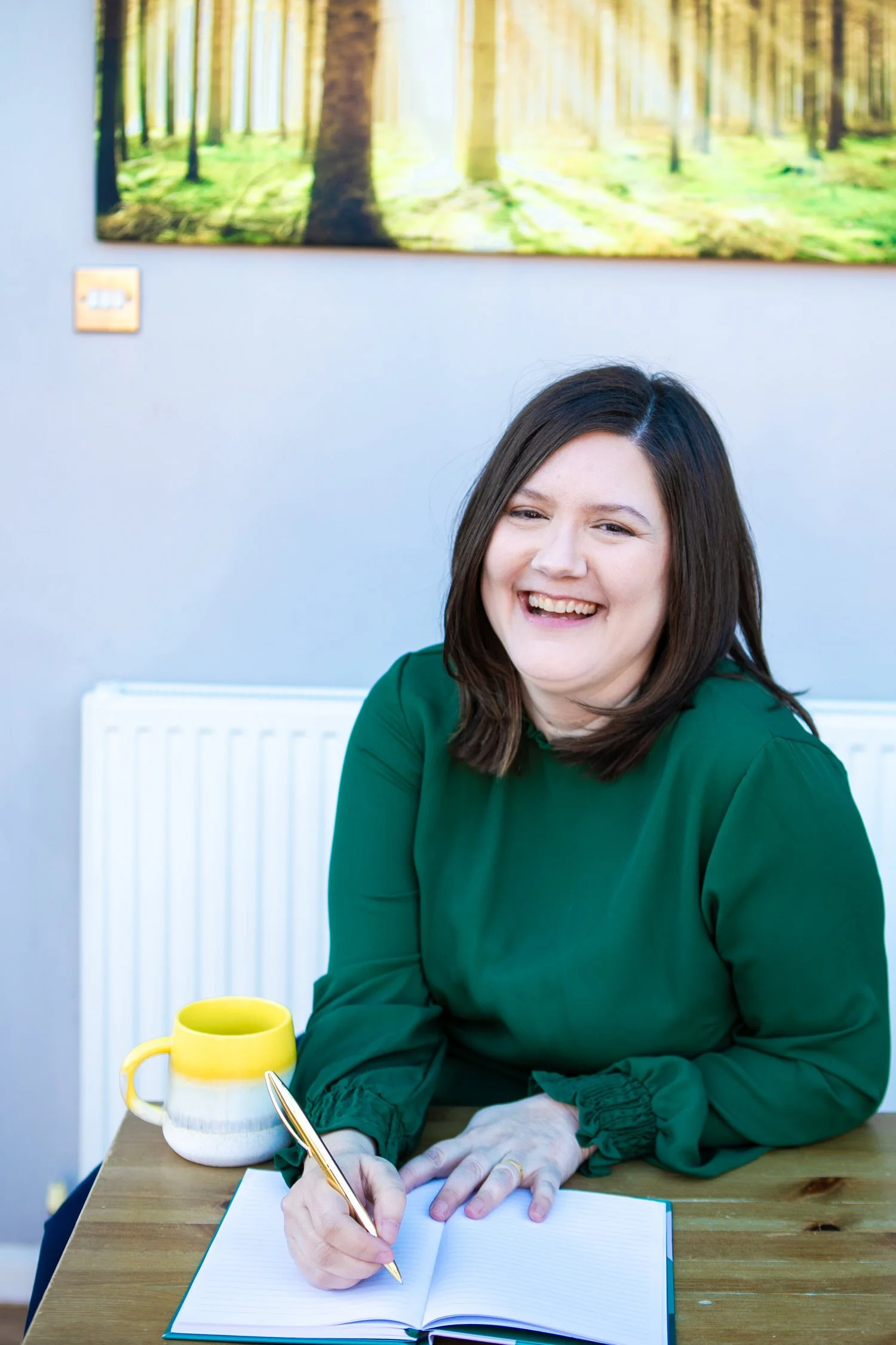 A woman with dark brown hair wearing a green top is sitting at a wooden table. She is smiling and writing in a lined notebook with a gold pen. On the table, there is a white and yellow mug and a yellow bowl. Behind her, on the wall, there's a large, blurred image of a forest with sunlight filtering through trees.