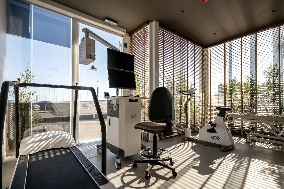 A modern physical therapy or rehabilitation room with large windows, sunlight, treadmill, exercise bike, chair, and medical equipment.