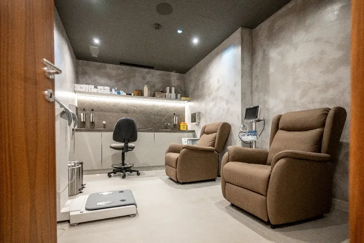 A medical room with two brown recliner chairs, a black office chair, a monitor on a stand, a small table, and medical supplies on shelves.