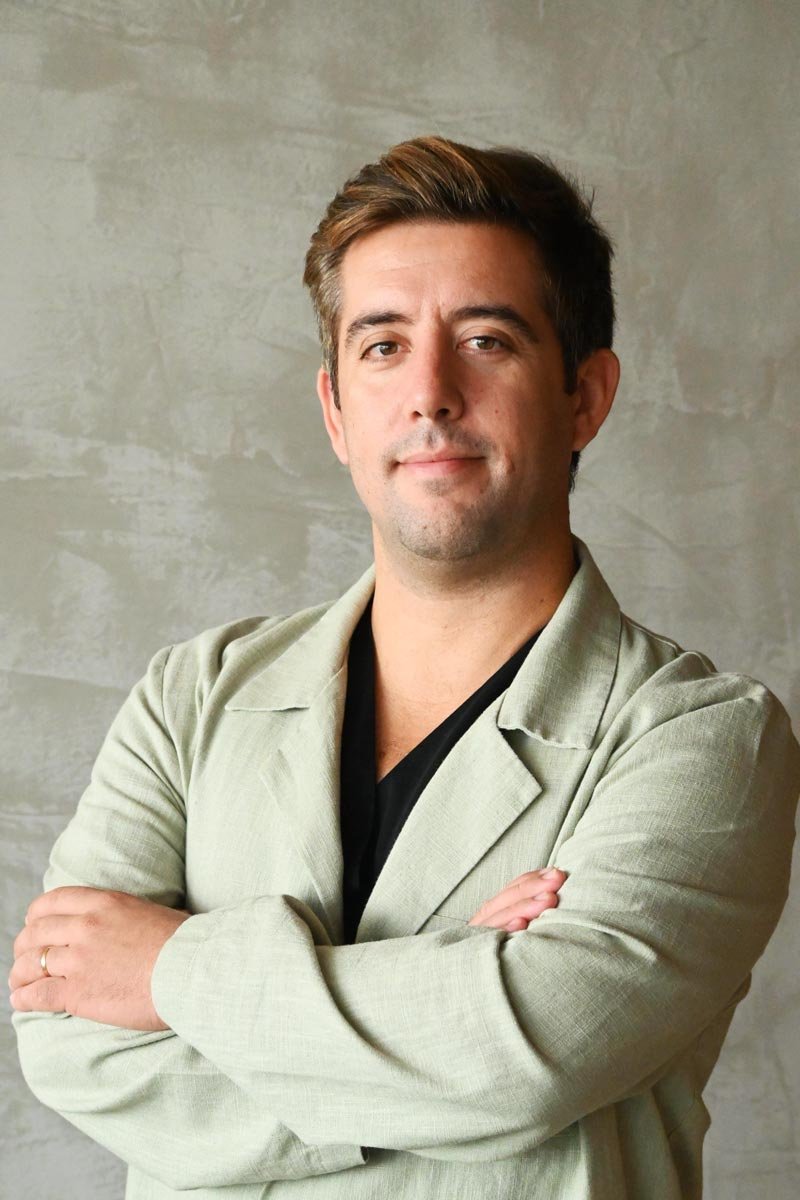 A young man with short brown hair and light skin, smiling and wearing a pink and white striped button-up shirt, standing indoors near a column with a blurred background.